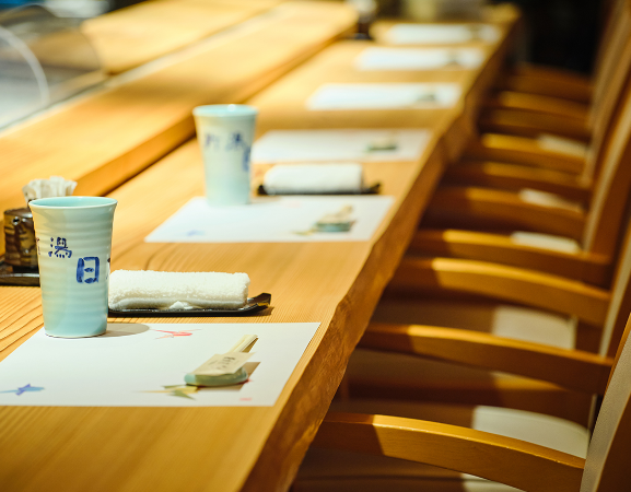 Chairs lined up at a wooden counter and seats with hot water, chopsticks, and white hand towels provided.