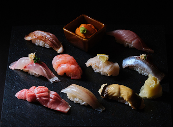 Various types of sushi beautifully arranged on a black plate and a square bowl with sea urchin in the center.