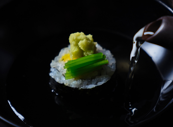 Sushi served in a black bowl with dashi being poured over it.Wasabi and green onions are on top.